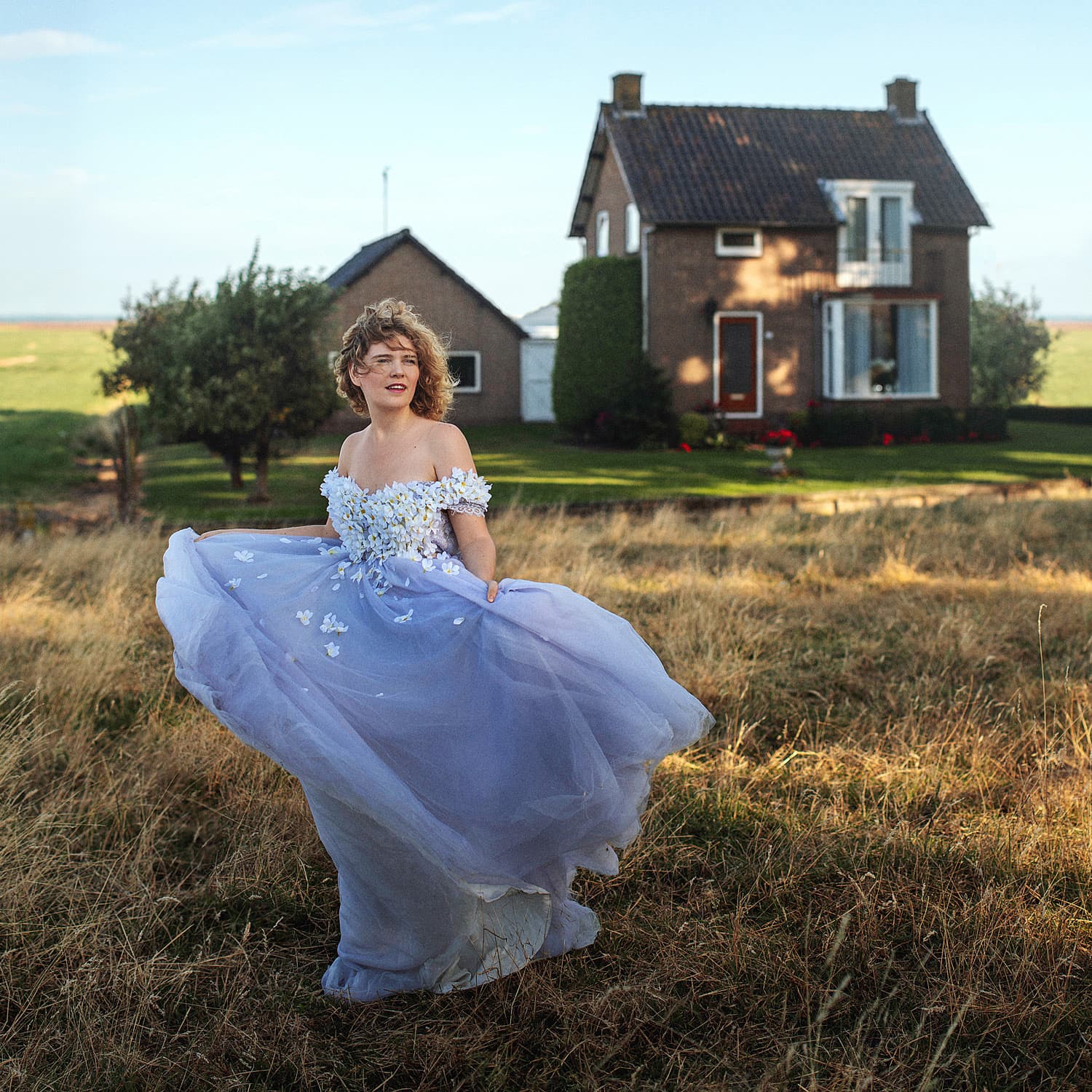 NinaLynn in een lavendelblauwe jurk in het gras voor een boerderij