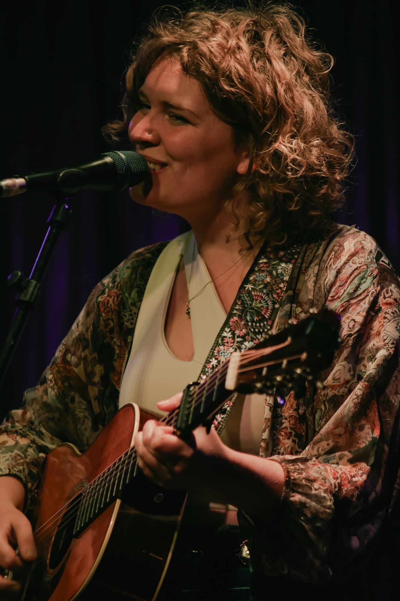 Nina zingt lachend met gitaar achter de microfoon