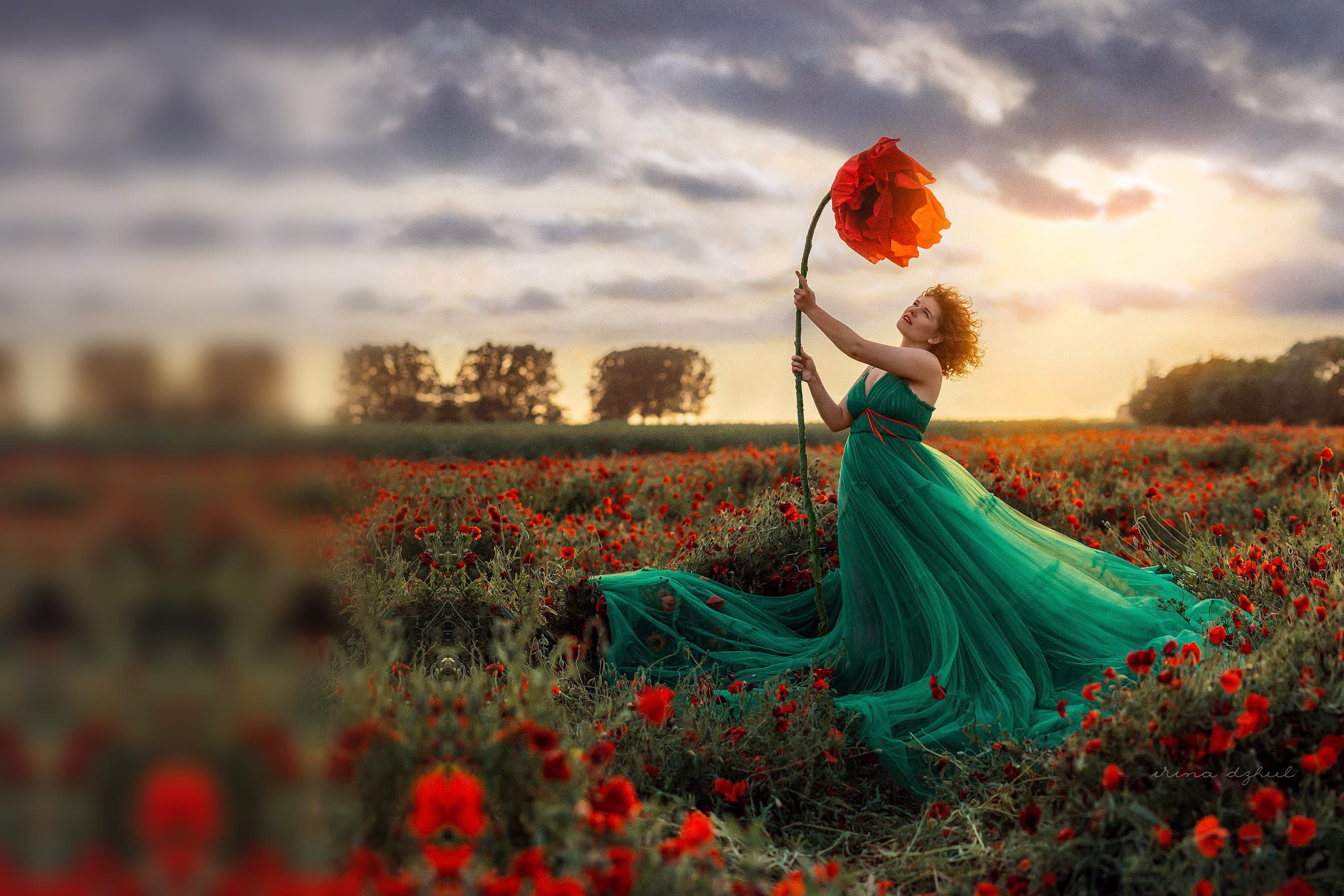 Nina in a green gown in a poppy field, holding up a giant poppy