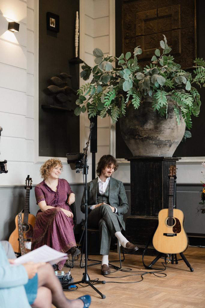 NinaLynn en een gitarist zitten lachend te luisteren tijdens een ceremonie, naast een grote vaas met takken