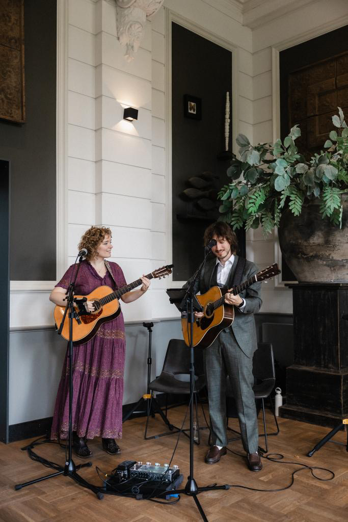 NinaLynn en een gitarist spelen samen op twee akoestische gitaren in een zaal met witte lambrisering
