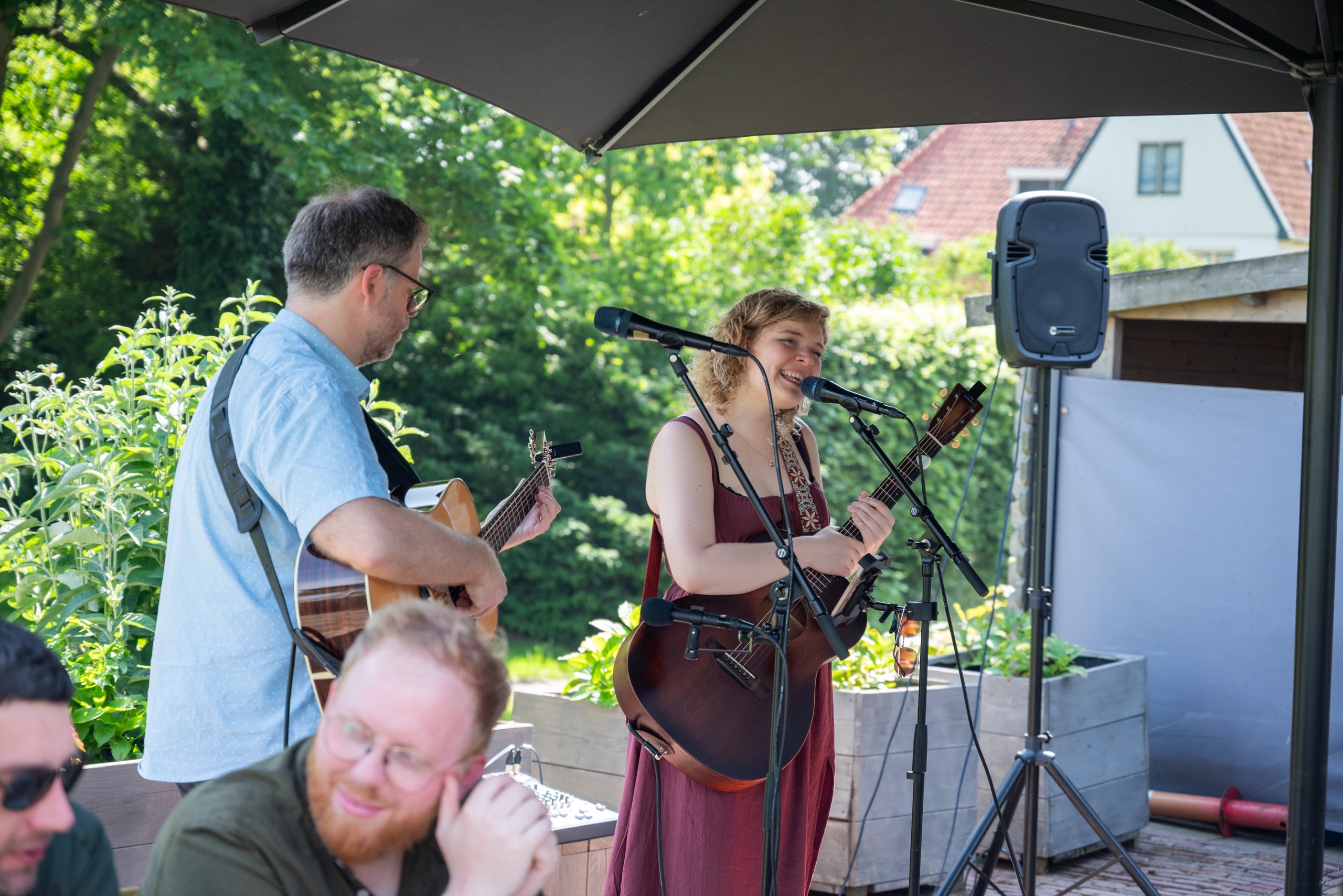 NinaLynn zingt lachend bij een microfoon, naast een gitarist, op een terras onder een parasol