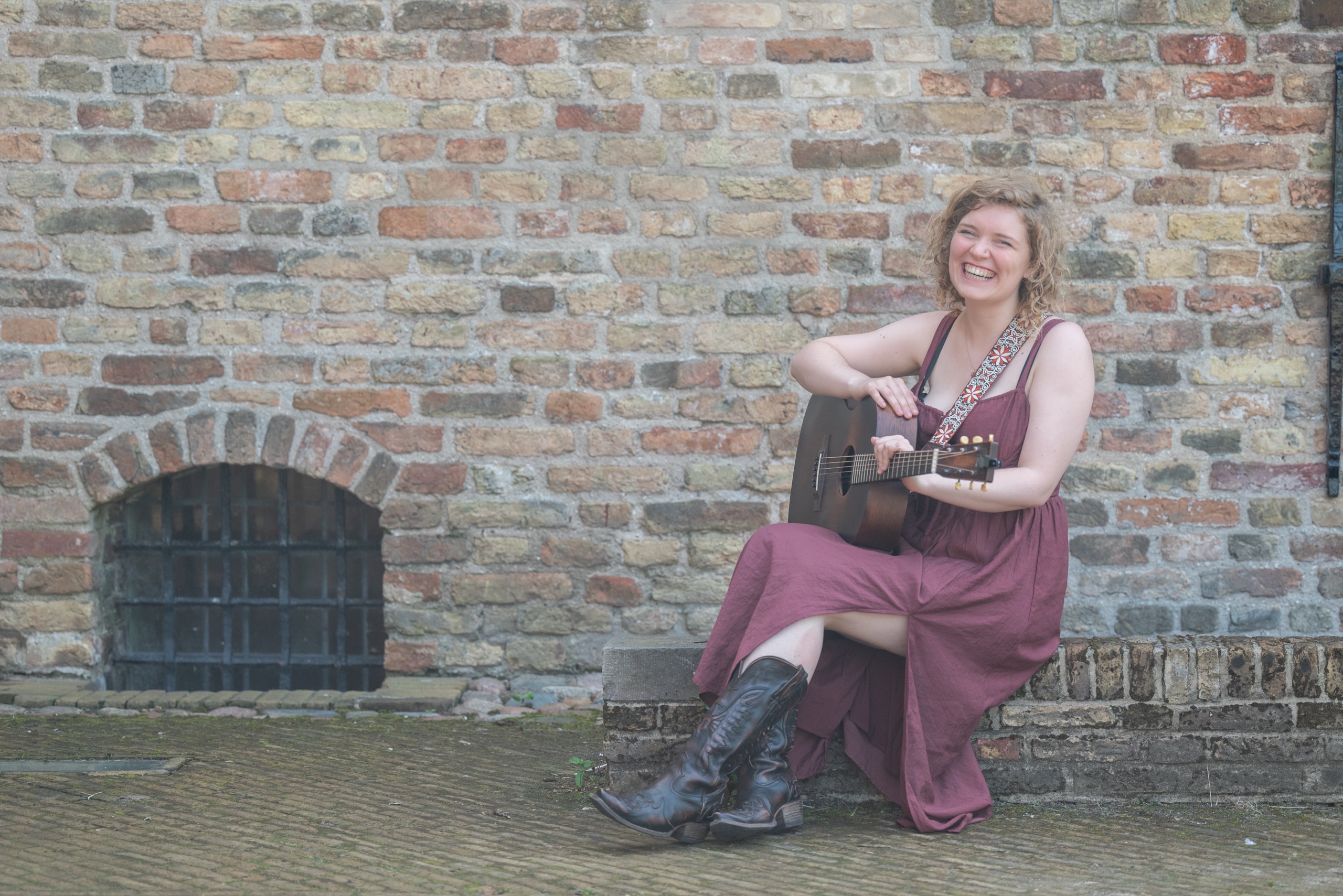 NinaLynn zit lachend met haar gitaar op een muurtje voor een bakstenen muur