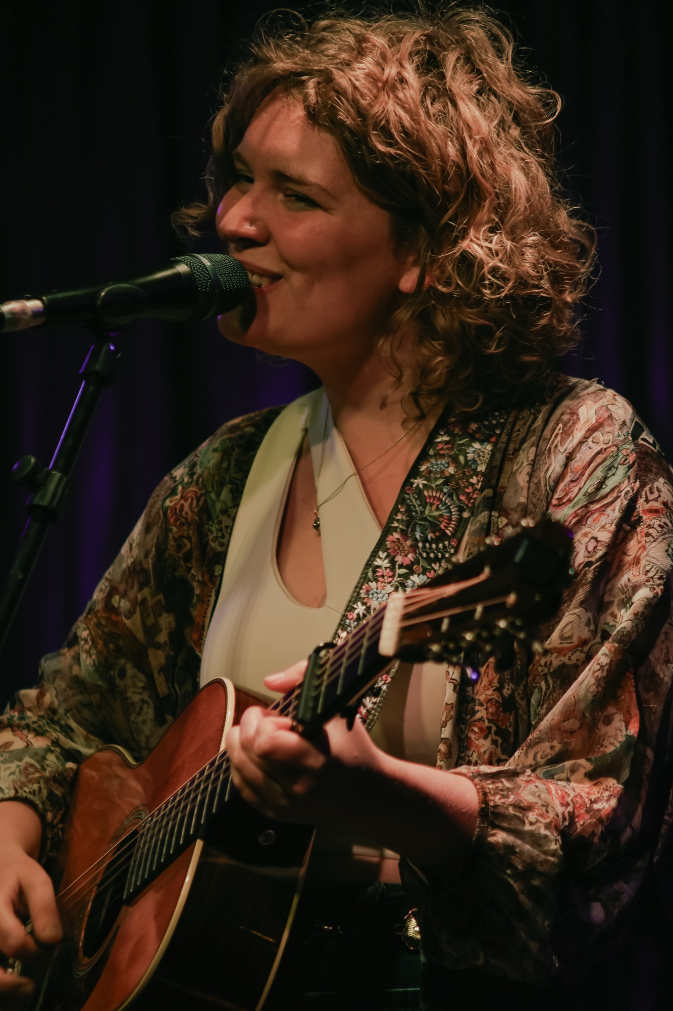 Nina zingt lachend met gitaar achter de microfoon