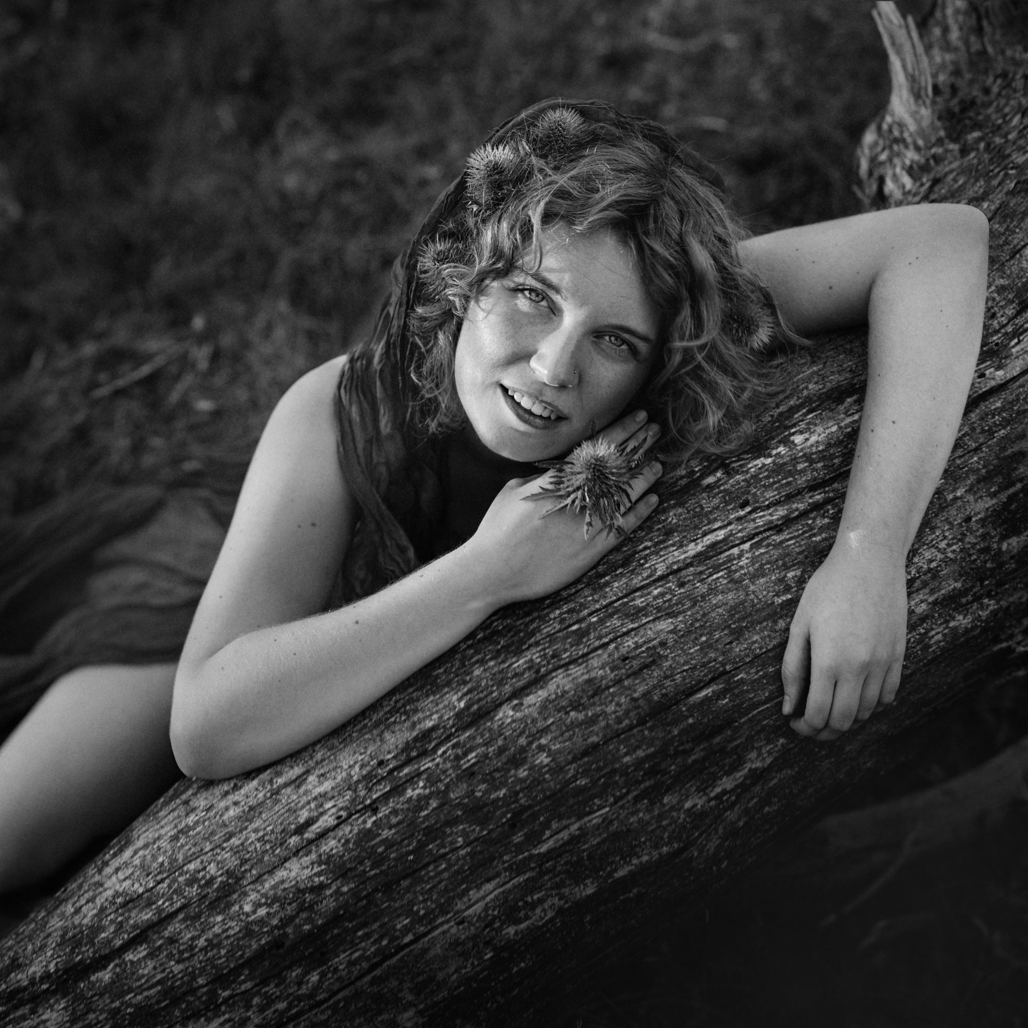 Nina in zwart-wit, met distels in haar haar en in haar hand, leunend op een gevallen boomstam
