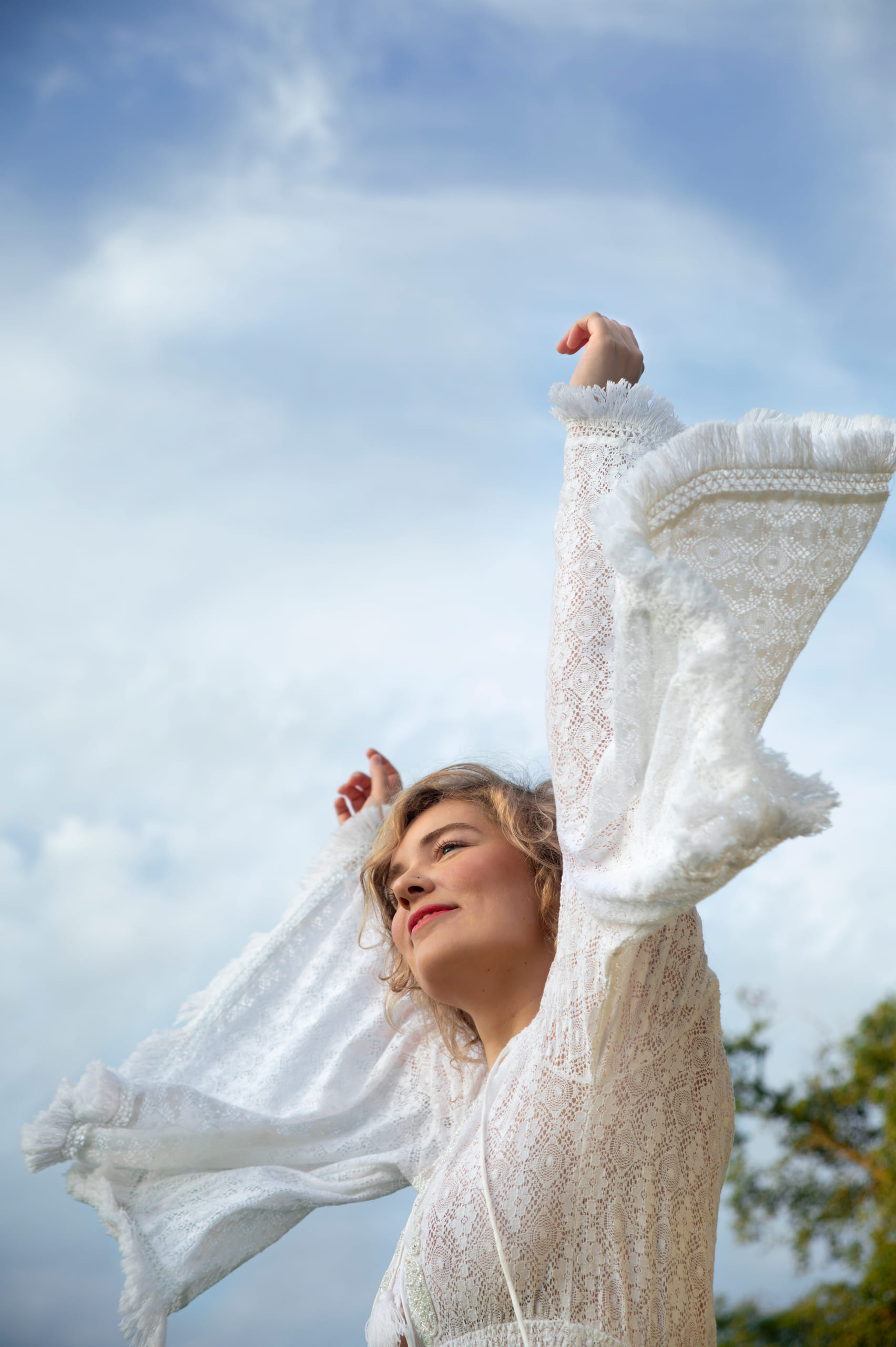 NinaLynn in een witte kanten blouse, met haar armen omhoog tegen een blauwe lucht