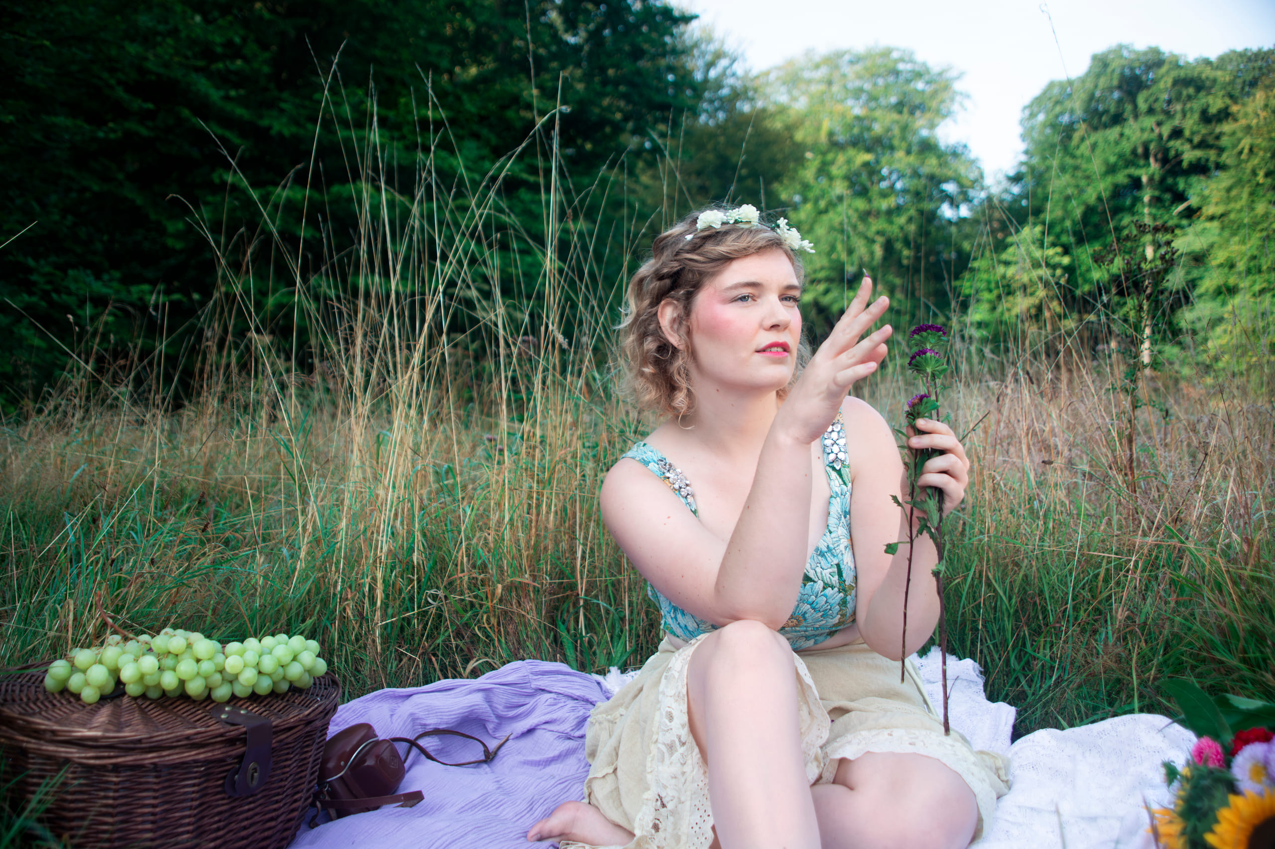 NinaLynn zit op een picknickkleed in hoog gras, met een bloemenkrans en een bosje asters in haar hand